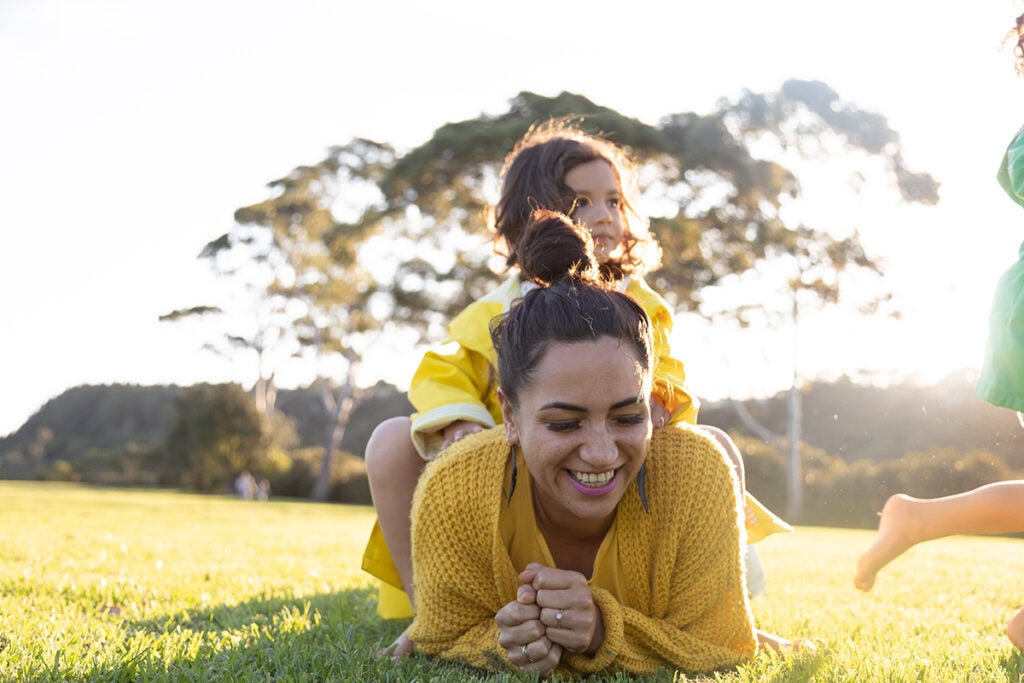 Mother laughing as one daughter is sitting on top of her and the other is running