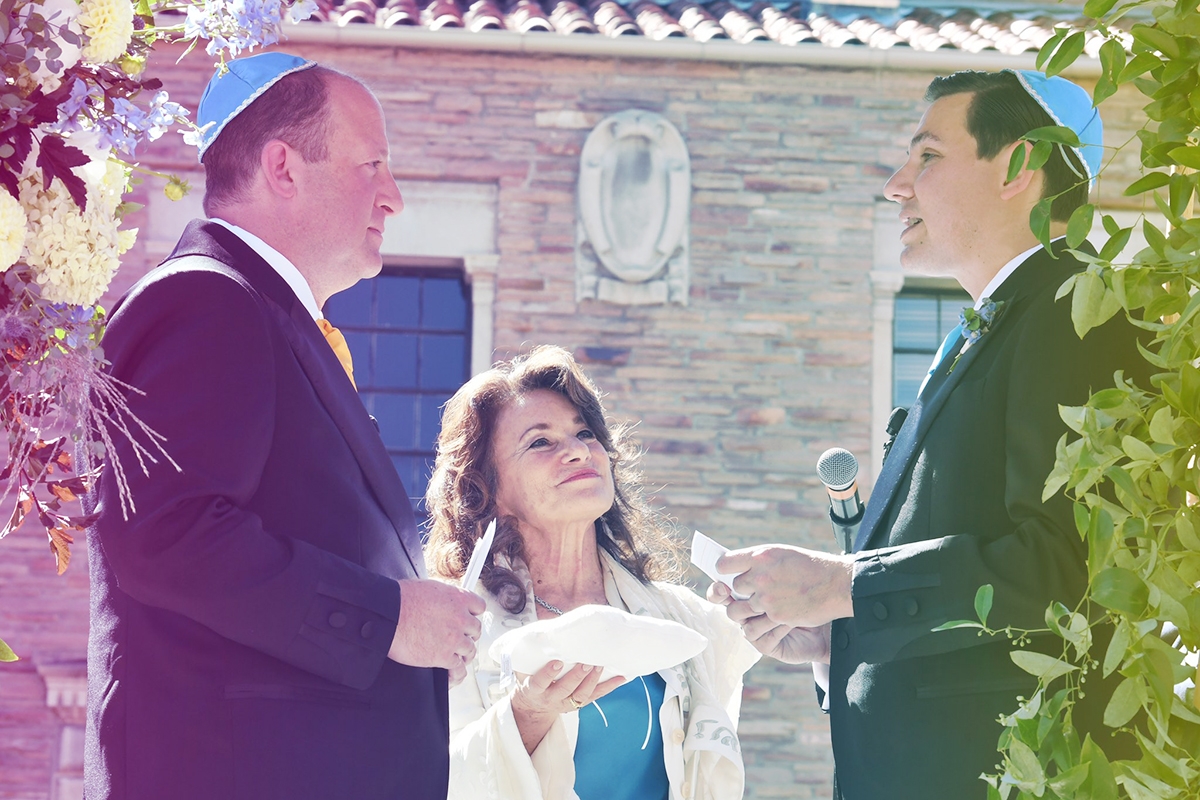 Jared Polis (L) marries partner Marlon Reiss (R) under a chuppah. Between them is officiating rabbi Tirzah Firestone