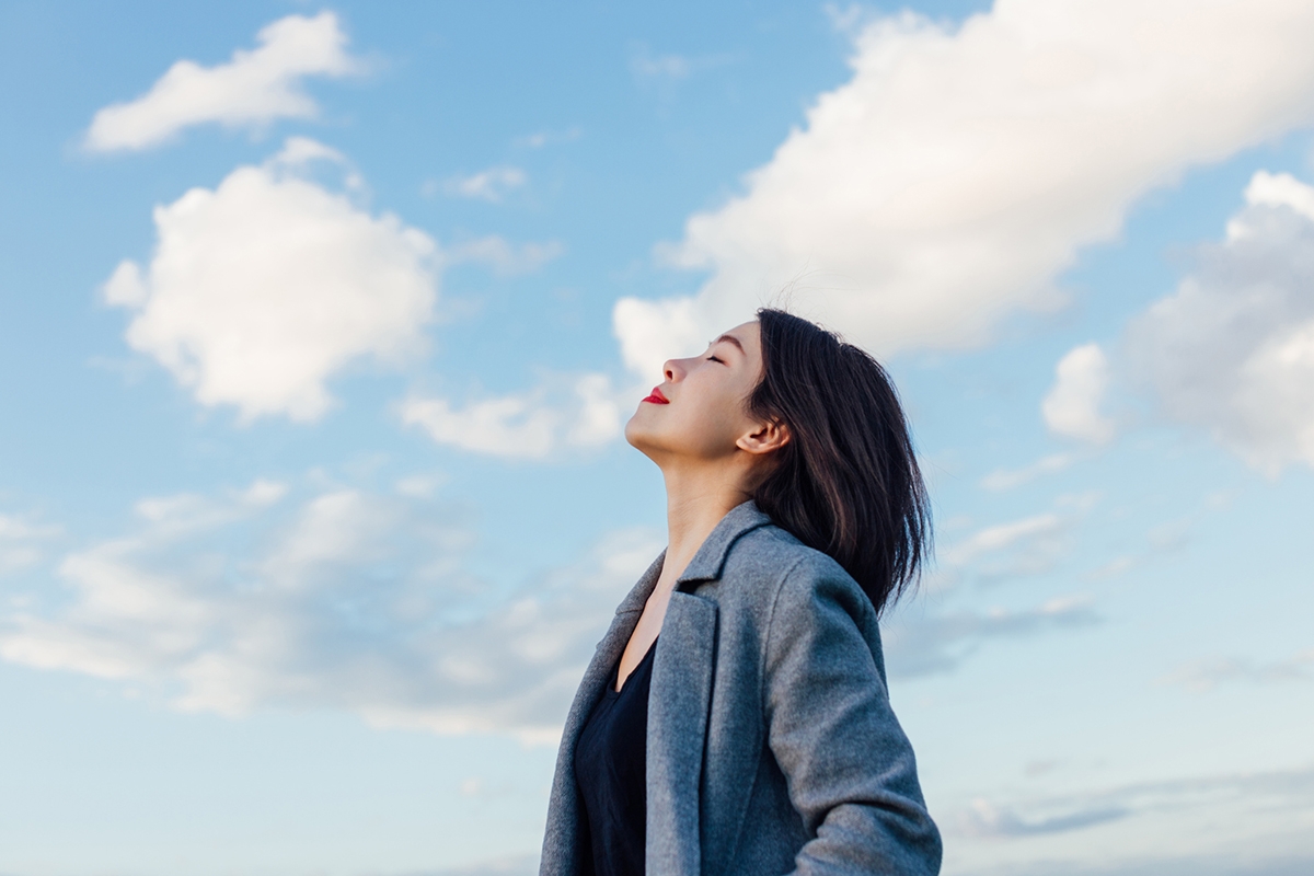 Portrait of positive young Asian woman with eye closed, enjoying sunlight under blue sky and clouds.