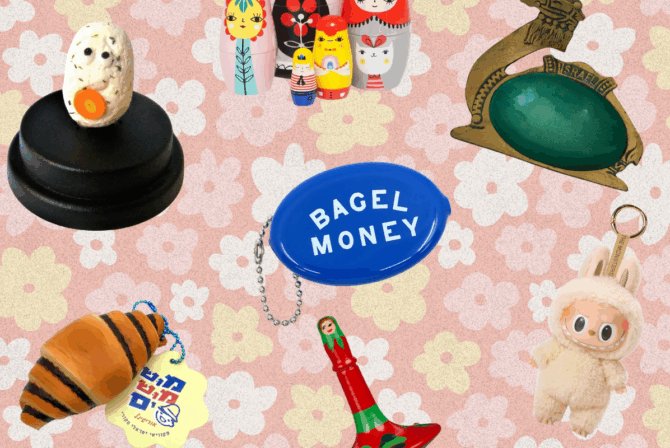 Multiple trinkets on a pink floral background. Trinkets include: a red dreidel, a cartoon gefilte fish sculpture, a set of nesting dolls, a brass and teal ashtray shaped like a camel, a rugelach keychain, a blue coin purse keychain that says "bagel money" and a Labubu.