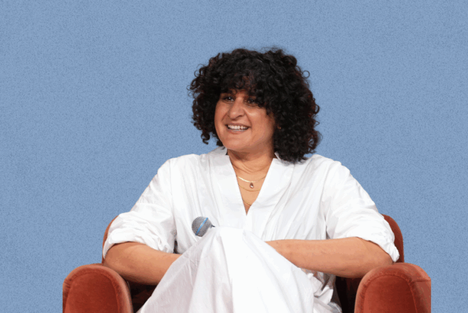 Samin Nosrat, seated and smiling while holding a microphone, against a blue background