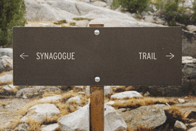 A sign on a hiking trail that has an arrow pointing one way toward "synagogue" and another pointing the other way toward "trail"